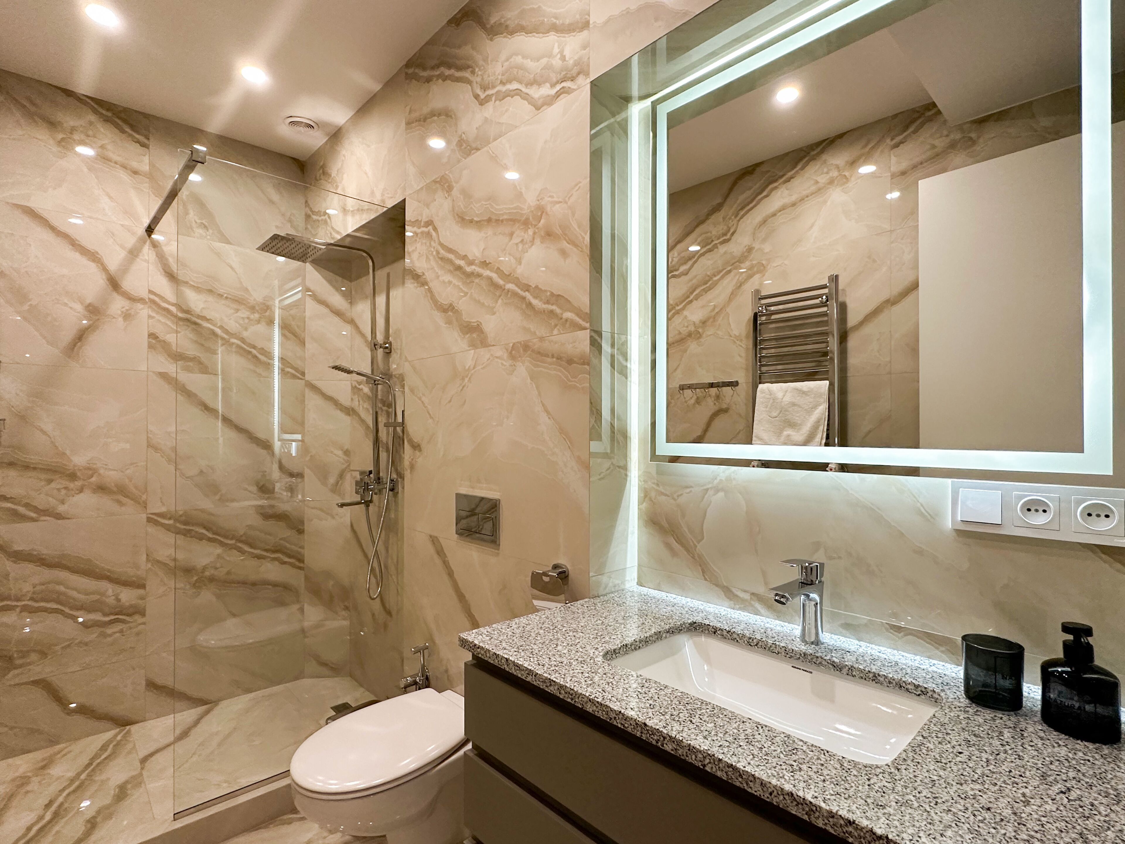 Een moderne badkamer met een douchecabine van glas, een wit toilet, een wastafel met een granieten blad en een grote spiegel.