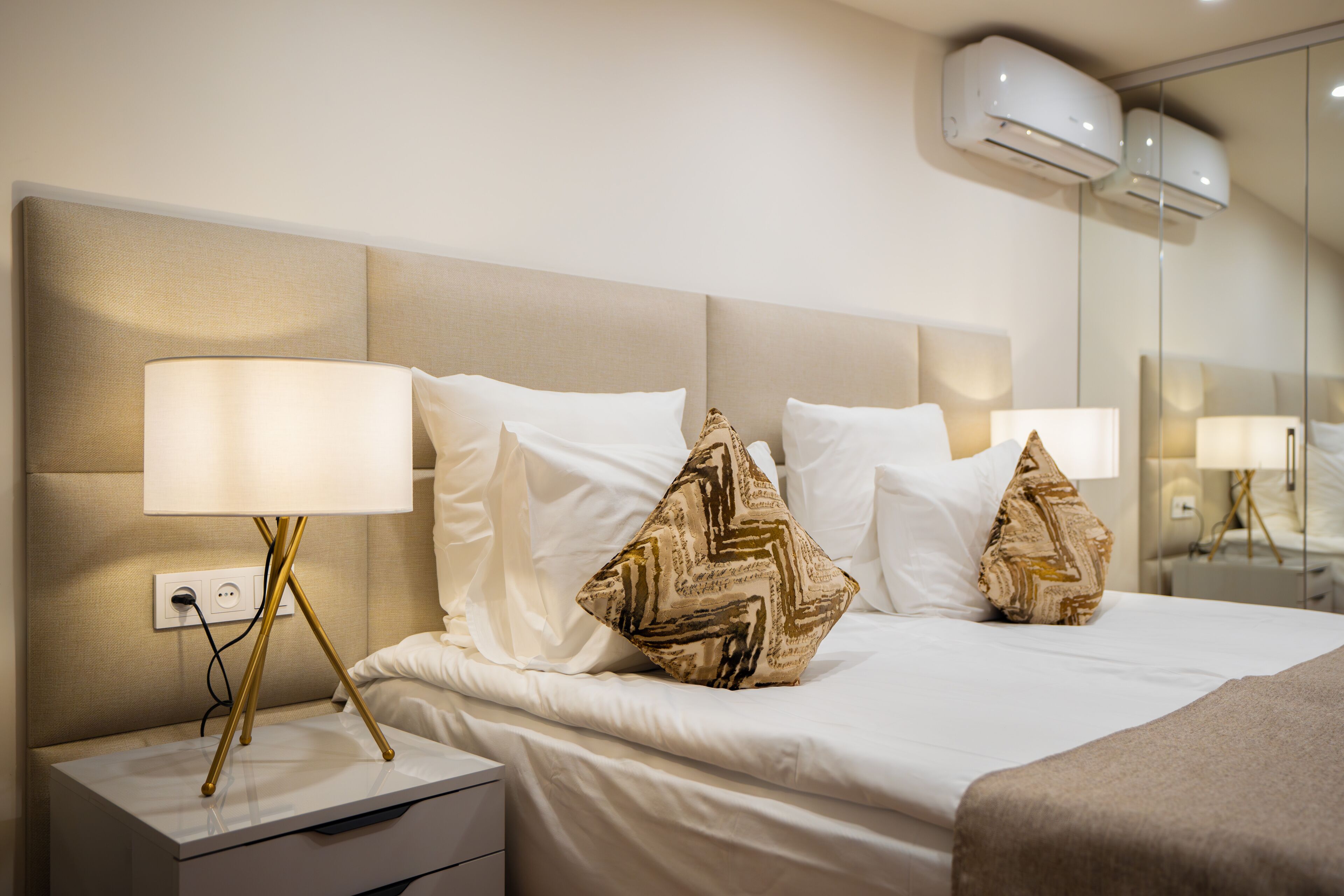 Una habitación de hotel con una cama grande, ropa de cama blanca y almohadas decorativas. Hay una mesita de noche con una lámpara y un enchufe.