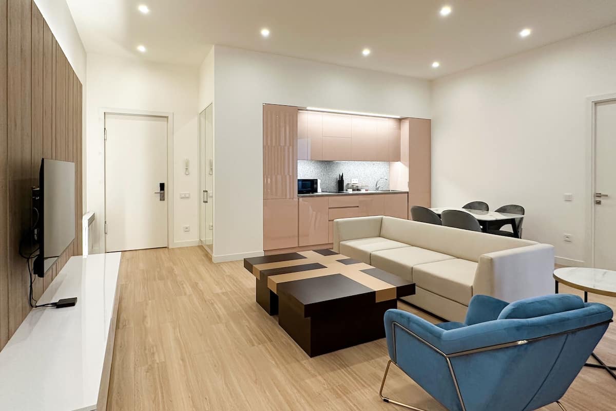 Un salon moderne avec un canapé, une table basse et un fauteuil bleu. On aperçoit une cuisine à l’arrière-plan, avec des placards et un plan de travail.