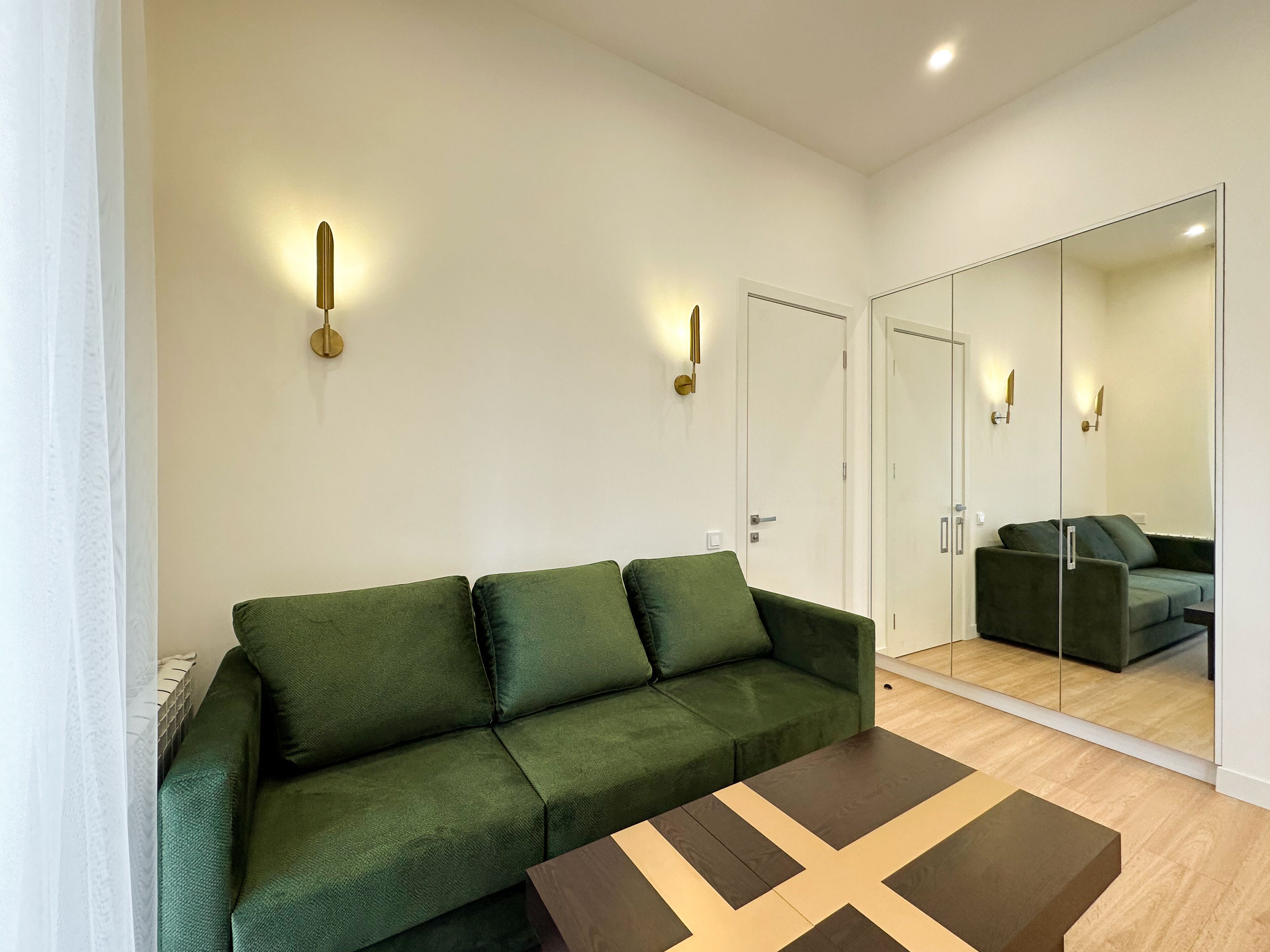 Un moderno salón con un sofá verde, una mesa de centro de madera y un espejo grande que refleja la habitación.