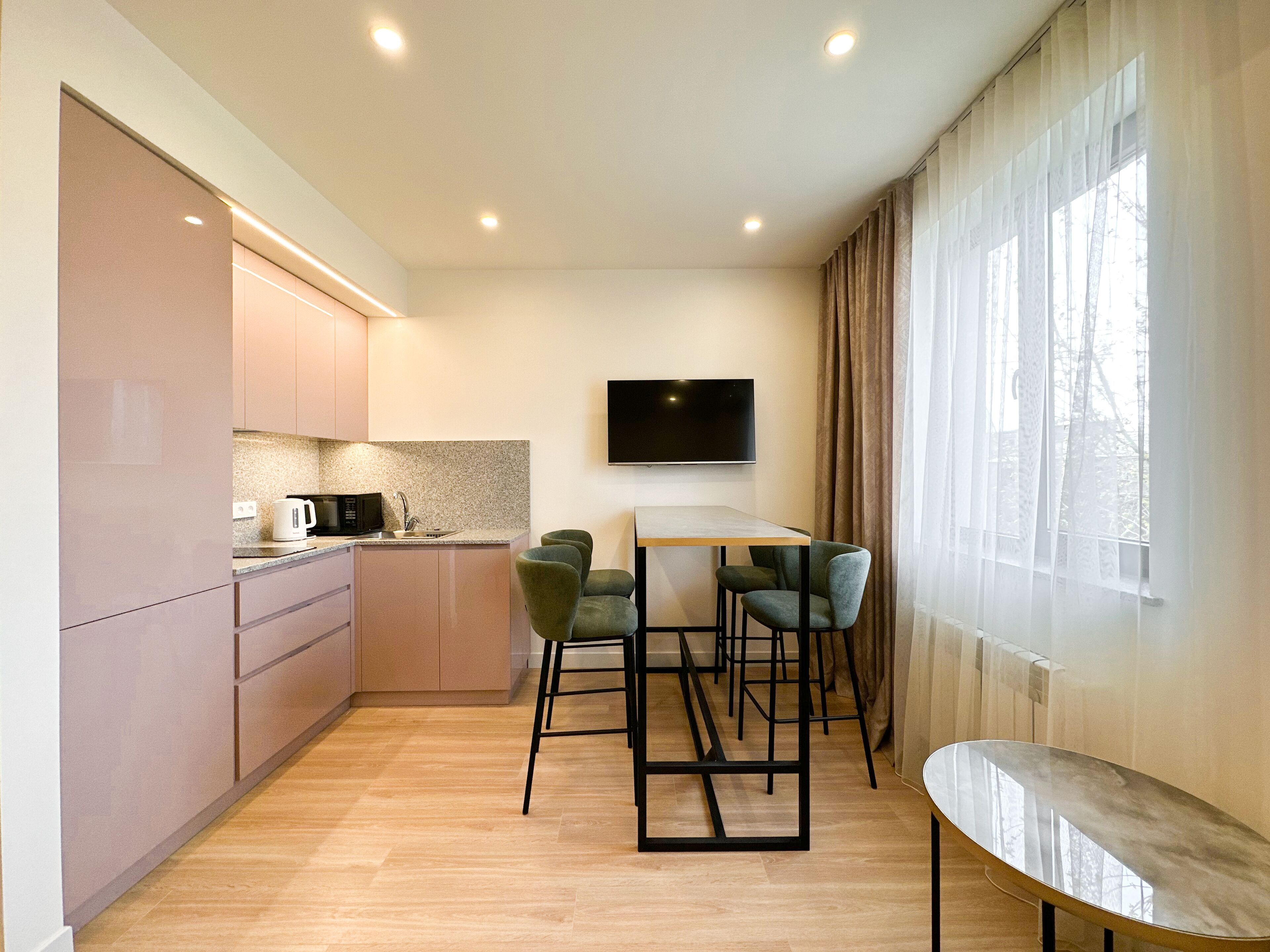 A modern kitchen with a built-in microwave, a coffee maker, and a sink. There is a dining area with a table and four chairs. A flat-screen TV is mounted on the wall above the table.