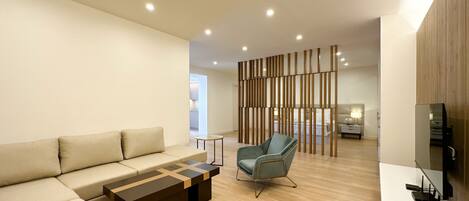 A modern living room with a sofa, a coffee table, and a chair. There is a wooden partition in the background, and a TV on the right side.
