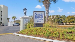 A sign for Sands Beach Club Resort with a parking lot and palm trees in the background. - Gorgeous views from this oceanfront condo in Shore Drive (Myrtle Beach)