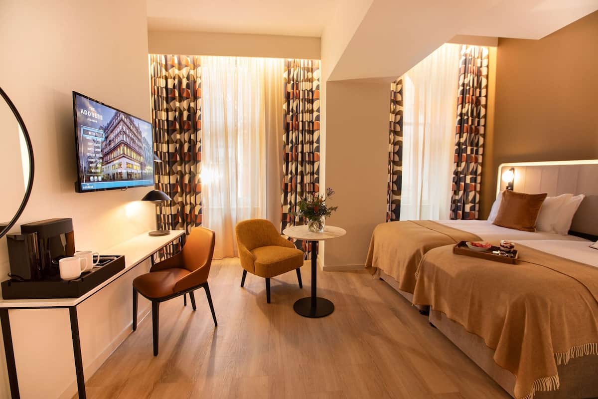 Ett hotellrum med en säng, ett skrivbord med en kaffemaskin, en stol, ett litet bord med en vas och gardiner.