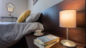Un lit bien fait, avec une couette grise et blanche, un coussin à motifs jaunes, une table de chevet sur laquelle se trouvent un livre, une tasse et une lampe.