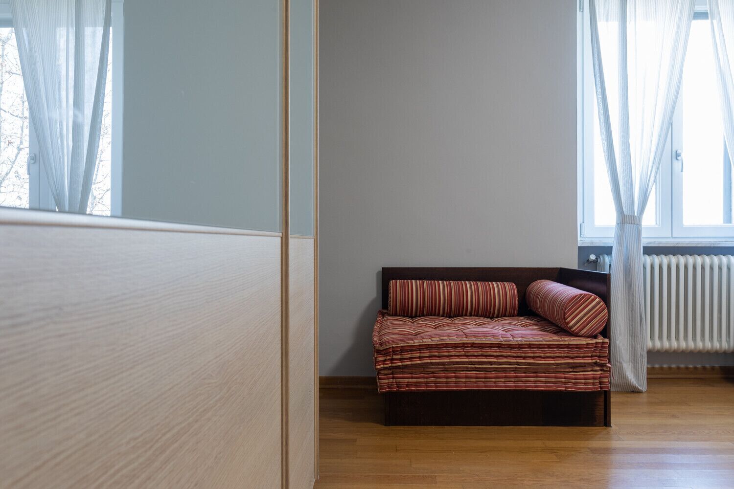 A room with a wooden floor, a bed with striped bedding, a window with sheer curtains, and a radiator.