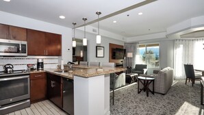 A modern kitchen with a built-in microwave, oven, and coffee maker. There is a dining area with a table and chairs, and a living room with a sofa and a TV.