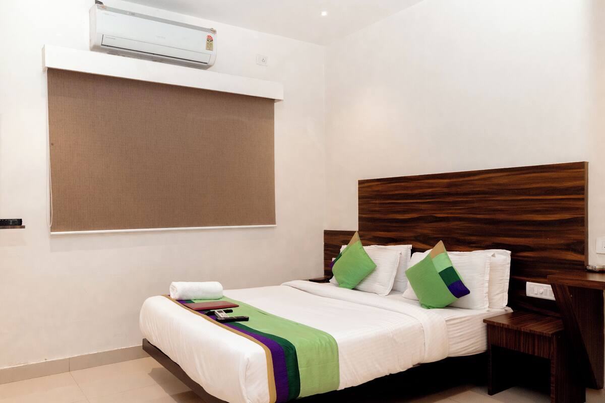 A hotel room with a bed, a nightstand, a wall-mounted air conditioner, and a window with a blind.