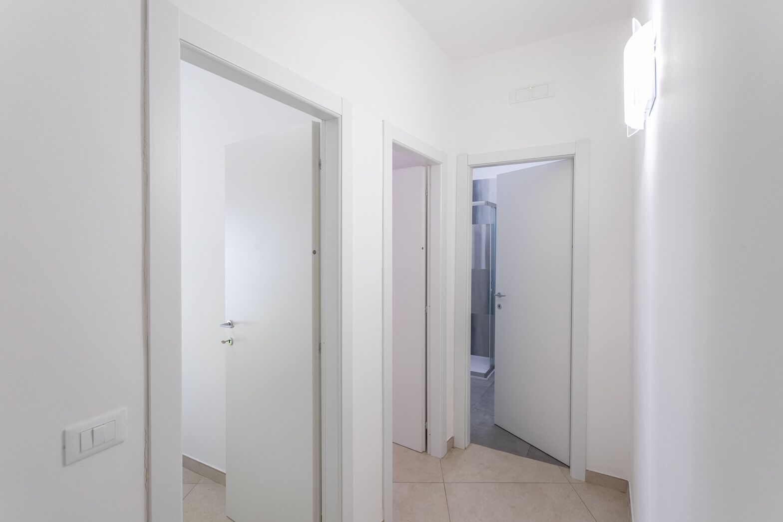 A hallway with white walls, a light switch, and a door leading to a bathroom.