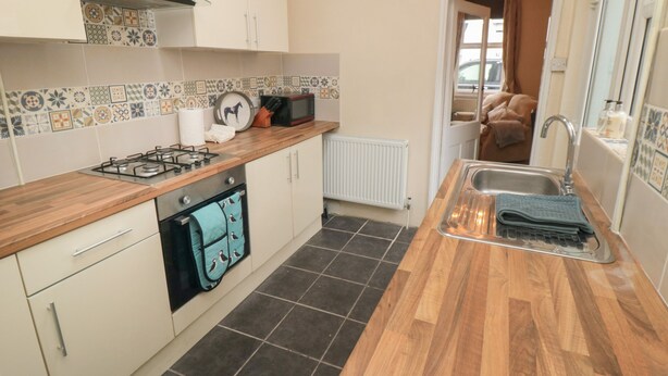 Une cuisine compacte équipée d’une cuisinière à gaz, d’un four et d’un four à micro-ondes, le tout installé sur un plan de travail en bois. Le mur derrière le plan de travail est recouvert de carreaux, et on peut apercevoir la pièce adjacente.