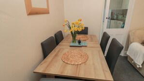 Un coin repas avec une table en bois, des chaises noires, des sets de table en fibres tressées et un vase rempli de fleurs jaunes.