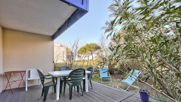 A wooden deck with a table and chairs, surrounded by greenery and a clear sky.