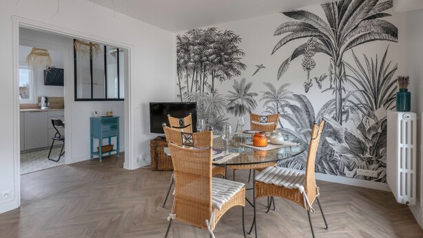 Uma área de refeições com mesa de vidro rodeada por cadeiras de vime, um mural de plantas tropicais na parede e uma televisão ao fundo.