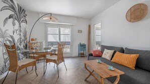 Uma sala de estar moderna com área de jantar, destacando uma mesa de vidro, cadeiras de vime, um sofá cinza e uma grande janela que proporciona iluminação natural.