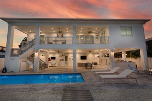 Una casa de dos pisos con piscina, mobiliario exterior y un balcón.