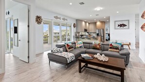 A modern living room with a grey sectional sofa, a wooden coffee table, and a view of the kitchen and outdoor patio. - Casa Del Mar~ Luxury Pool Home w/ Kayaks and Bikes! (Marathon)