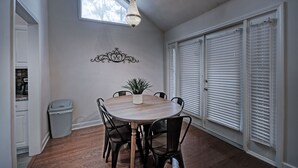 A dining area with a round table, six chairs, a potted plant, a chandelier, and a window with blinds.