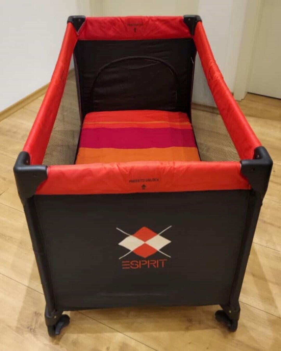 A black and red portable crib with a mattress, labeled "ESPRIT," on a wooden floor.