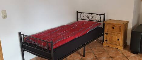 A single bed with a metal frame and a red bedspread, a wooden nightstand with drawers, and a black chair.