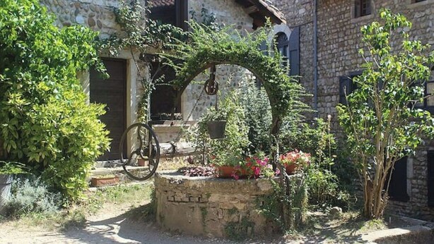 Un bâtiment rustique en pierre, avec un vélo appuyé contre un mur de pierre, entouré de verdure et de plantes en fleurs.