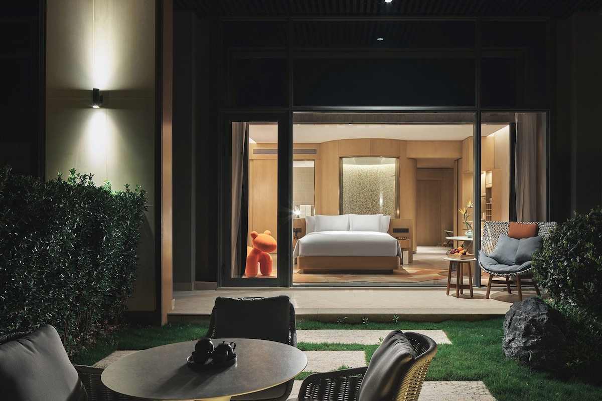 Une chambre d’hôtel moderne dotée d’un grand lit, d’un coin salon avec un canapé et une table, et offrant une vue sur la pièce grâce à une porte vitrée.