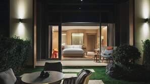 Quarto de hotel moderno com uma cama grande, área de estar com um sofá e uma mesa, e vista do quarto através de uma porta de vidro.