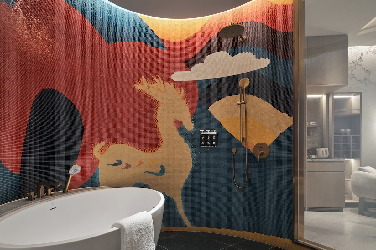 Une salle de bain dotée d’un grand mur décoré d’une mosaïque colorée, d’une baignoire sur pieds et d’une douche séparée par des parois en verre.