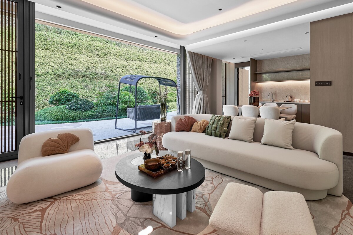 A modern living room with a sofa, armchairs, a coffee table, and a view of a garden through large windows.