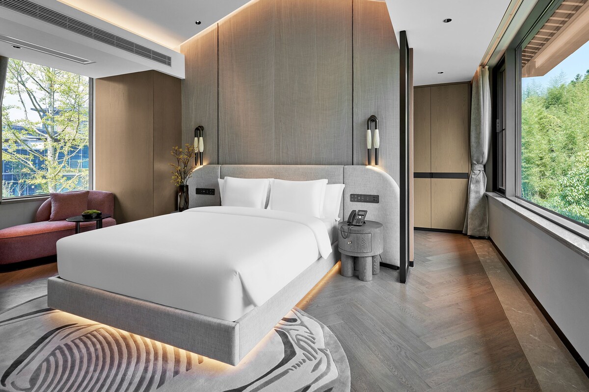 A modern hotel room with a large bed, bedside tables, a view of greenery, and a window with curtains.