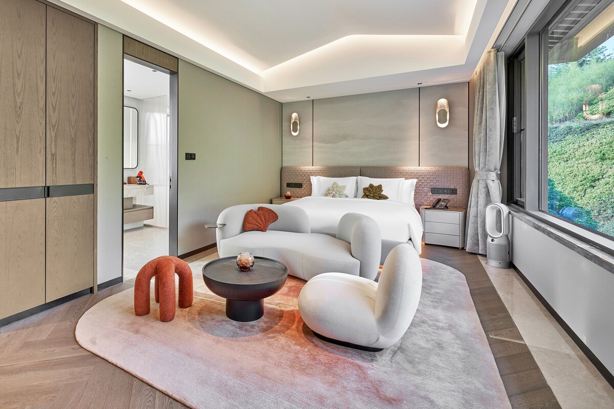 A modern hotel room with a large bed, a sitting area with a coffee table, a side table, and a view of greenery through the window.