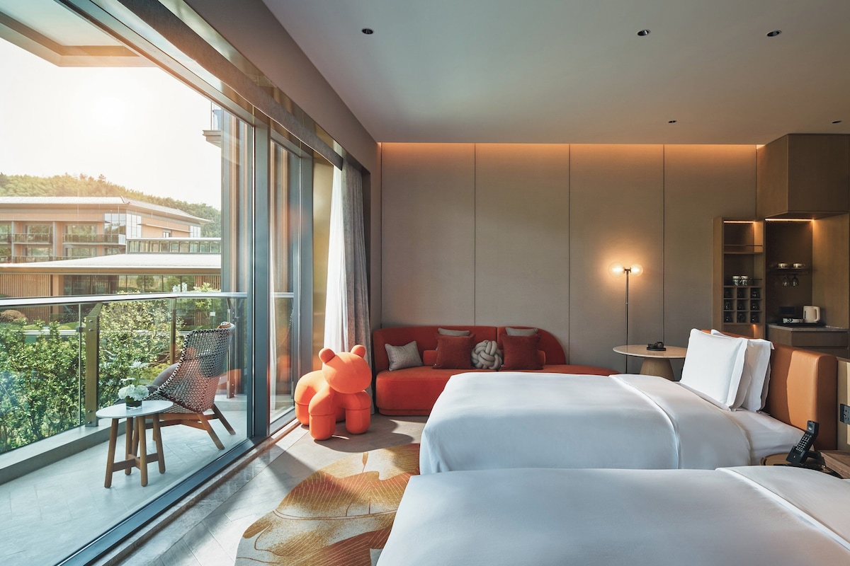 Une chambre d’hôtel moderne avec deux lits, un canapé rouge, une grande fenêtre et un balcon.