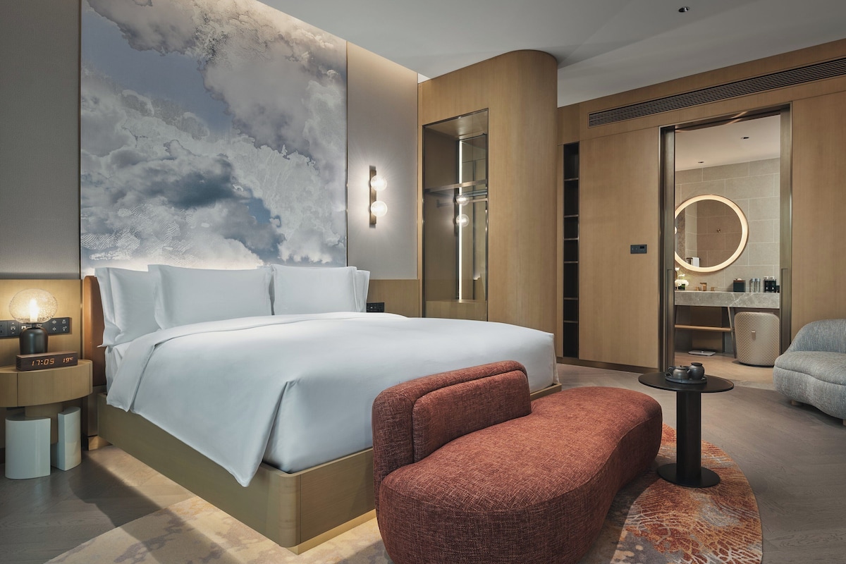 Une chambre d’hôtel moderne avec un grand lit, un coin salon comprenant un fauteuil rouge et une petite table, et une salle de bain visible à travers une porte ouverte.