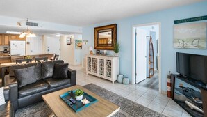 Una sala de estar moderna con un sofá de cuero negro, una mesa de centro de madera y un televisor. Hay una zona de cocina con un refrigerador y armarios, y un comedor con una mesa y sillas.