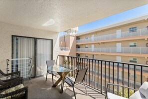 Un balcón con una mesa de vidrio y sillas de mimbre, con vistas a un edificio de varios pisos con balcones.