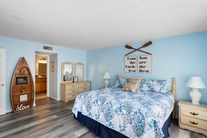 Un dormitorio con cama, dos mesitas de noche, cómoda y tocador con espejos. Hay almohadas decorativas y un adorno de pared con un mensaje inspirador.