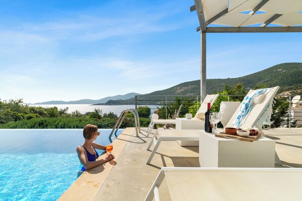 Un espace aménagé au bord de la piscine, avec des chaises longues, une table sur laquelle se trouvent une bouteille et un verre, et une vue sur la mer et les collines.