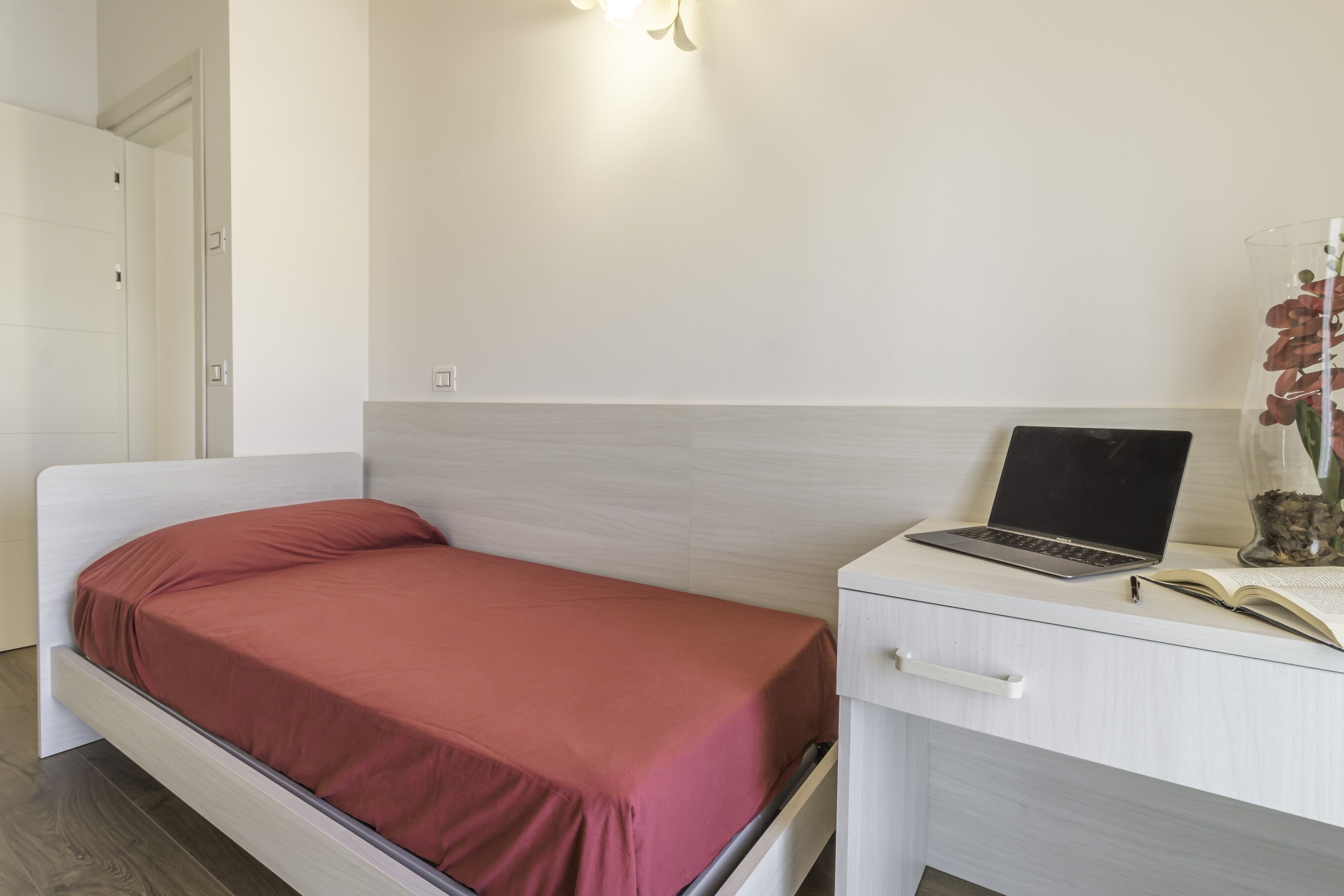 A bedroom with a bed, a desk with a laptop, and a vase with flowers.