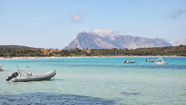 A coastal scene with clear turquoise waters, boats, and a mountain in the background. - Villa Asta (San Teodoro)