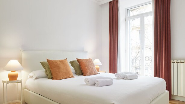 A neatly made bed with white linens, two bedside lamps, and a view of the outdoors through a window with red curtains. - Catedral | The Rentals Collection (San Sebastián)