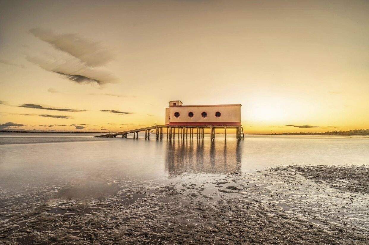 Un edificio de techo plano y cuatro ventanas circulares sobre un muelle que se adentra en una masa de agua tranquila durante la puesta de sol.