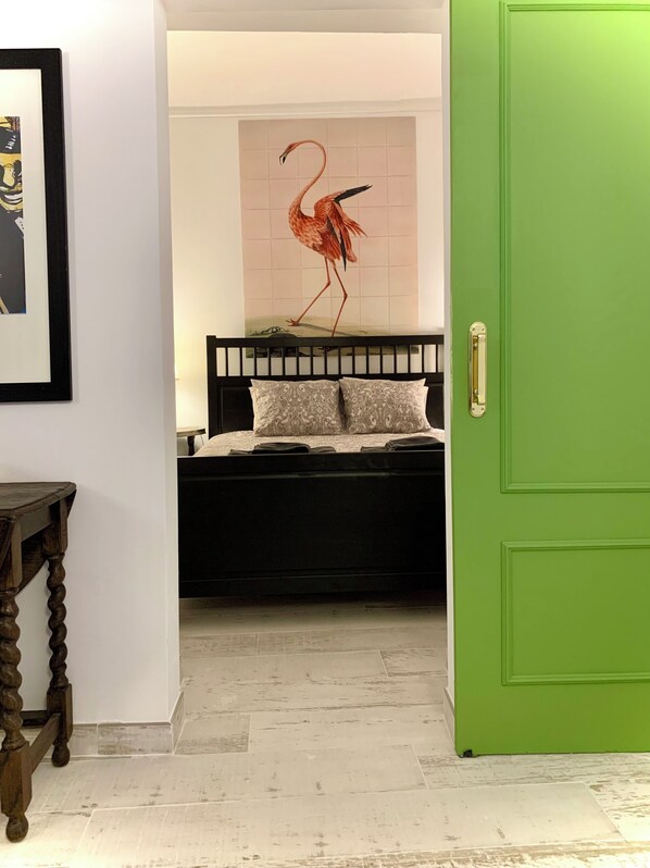A room with a green door, a black bed with patterned pillows, a wooden table, and a wall with a red bird painting.