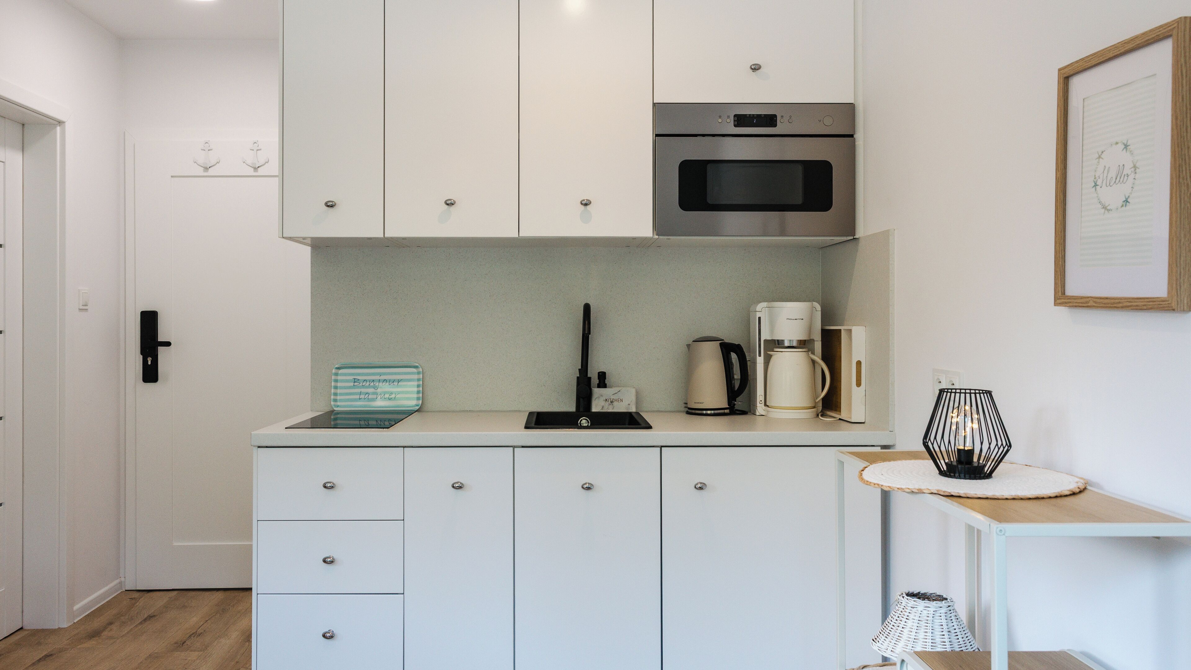 A compact kitchen with white cabinets, a black faucet, a coffee maker, and a framed welcome sign.