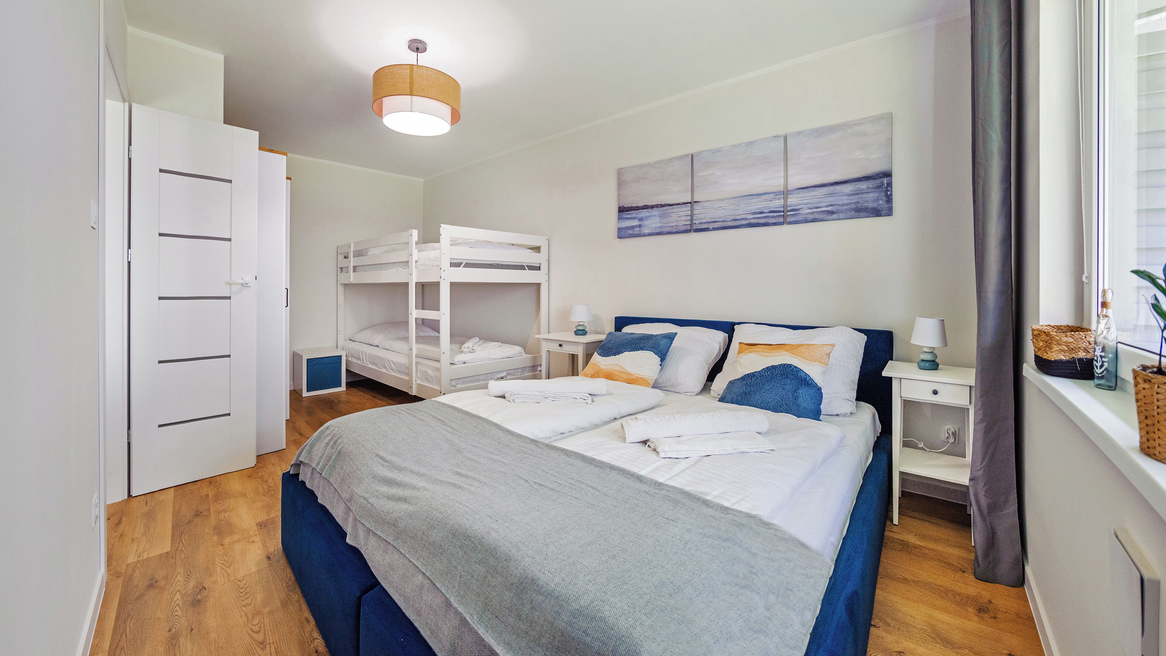 A bedroom with a bunk bed, a bedside table with a lamp, a wall-mounted painting, and a window with curtains.