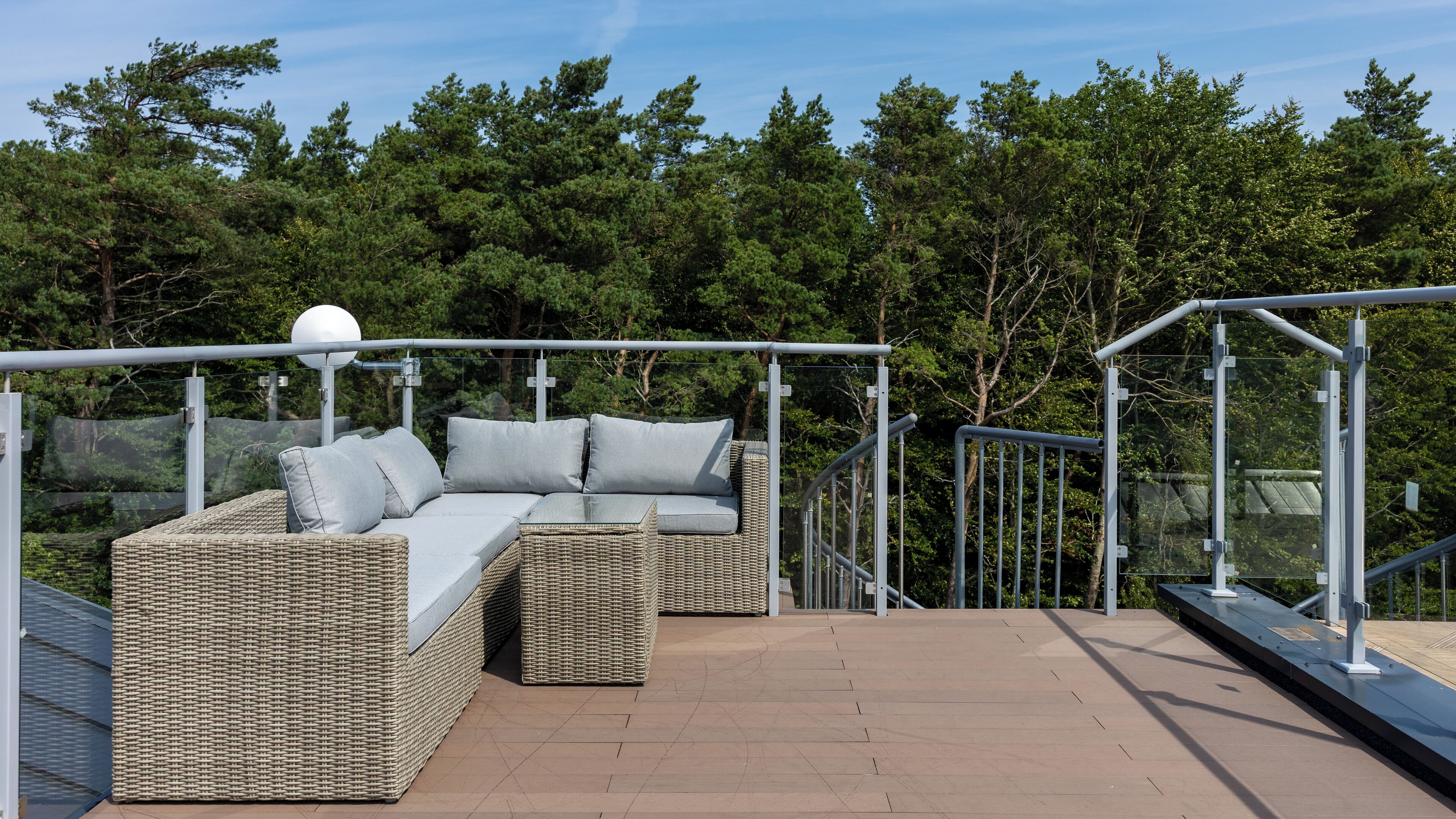 A wicker sofa with cushions on an outdoor deck with a railing and trees in the background.