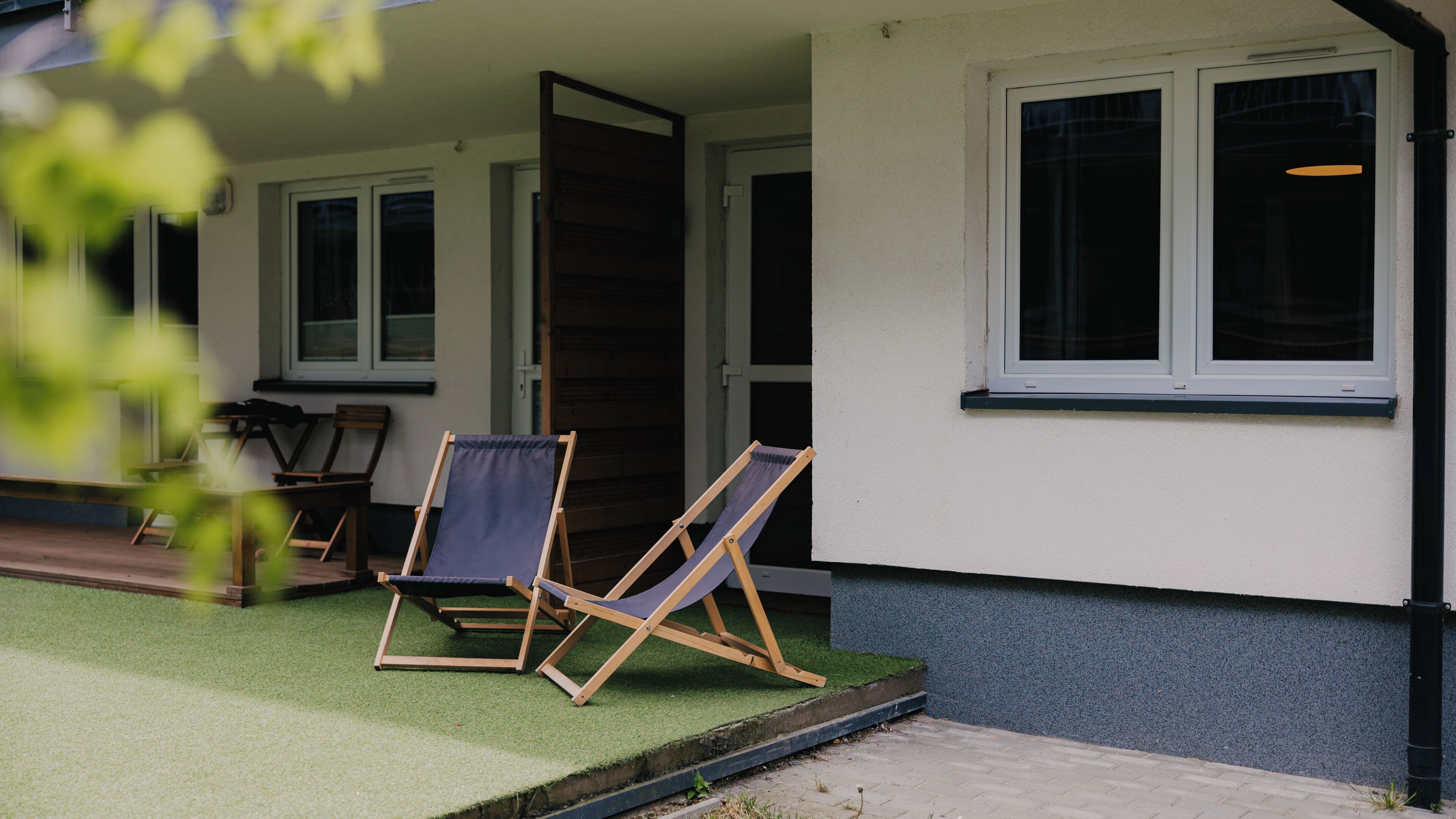 Un espacio al aire libre cubierto con dos sillas de madera y una mesa, una puerta y una ventana.