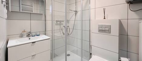 A modern bathroom with a glass shower enclosure, a white sink with two glass cups, and a toilet with a paper towel dispenser.