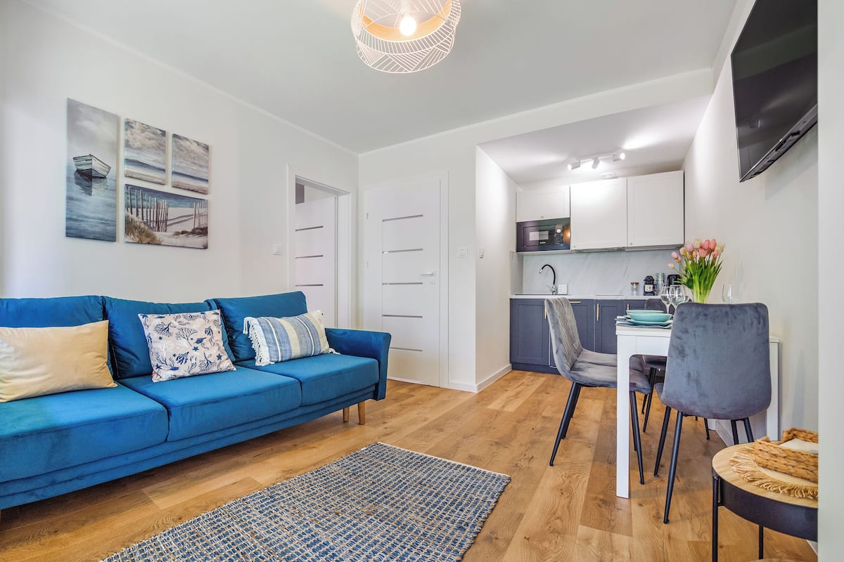 Ein modernes Wohnzimmer mit einem blauen Sofa, einem Essbereich mit Tisch und Stühlen, einer Küche mit weißen Schränken und einem an der Wand befestigten Flachbildfernseher.
