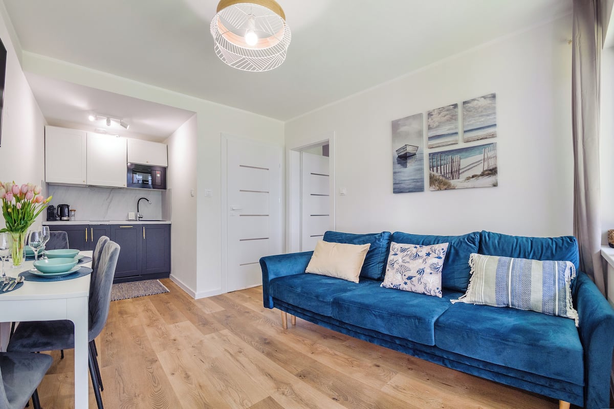 Ein modernes Wohnzimmer mit einem blauen Sofa, einem Esstisch mit Stühlen und einer Küchenzeile im Hintergrund.