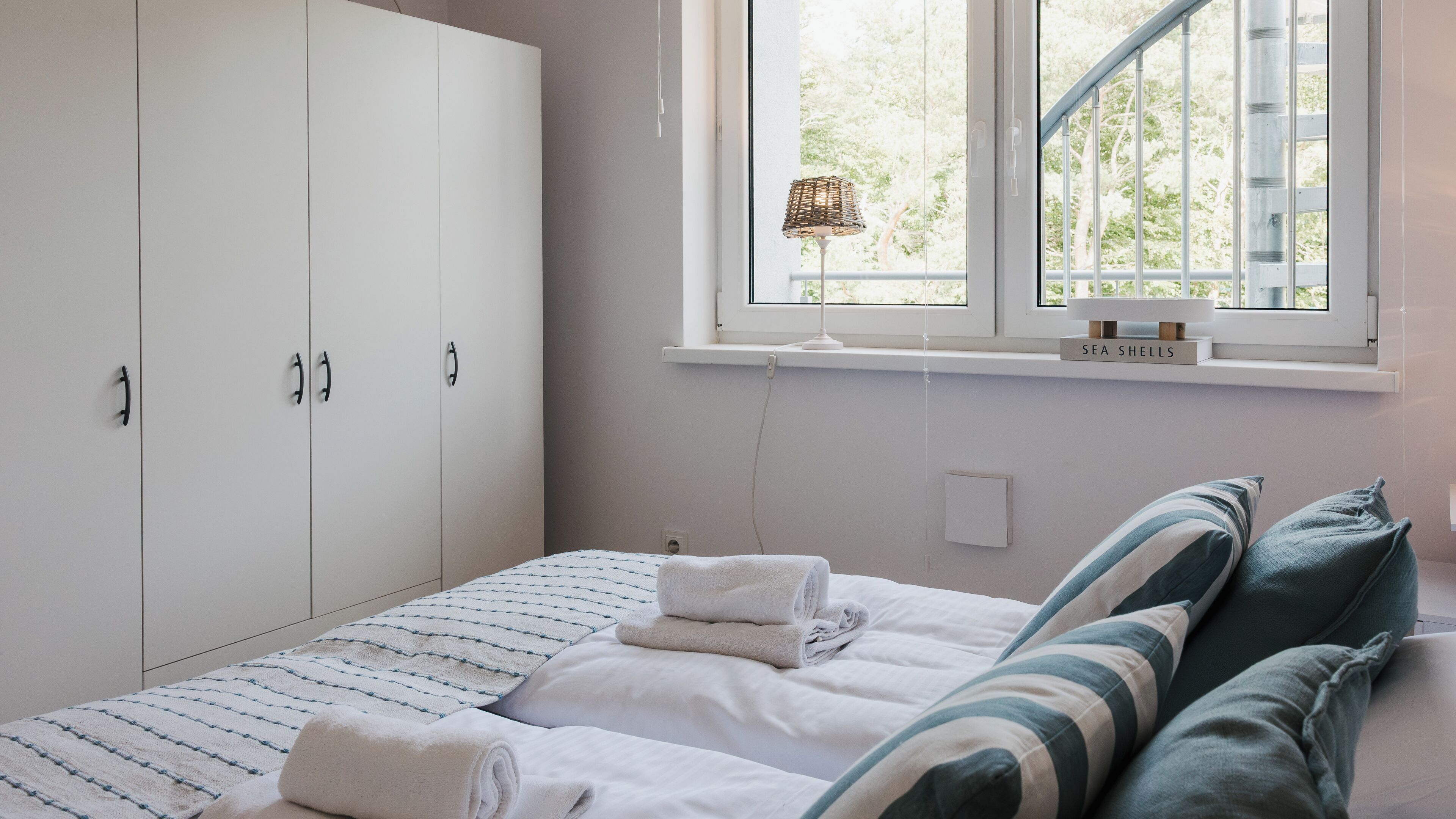 A bedroom with a bed, a window, a wardrobe, and a bedside table with a lamp.