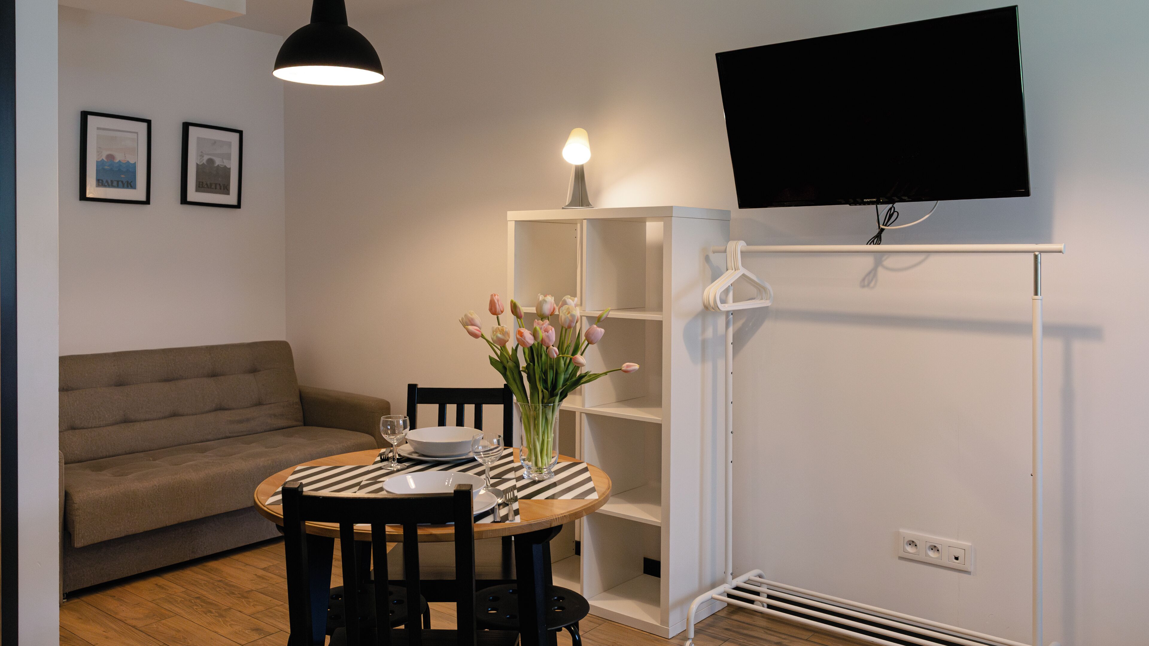 Una habitación pequeña y moderna con una mesa de comedor redonda, un sofá, un televisor montado en la pared y un estante blanco.